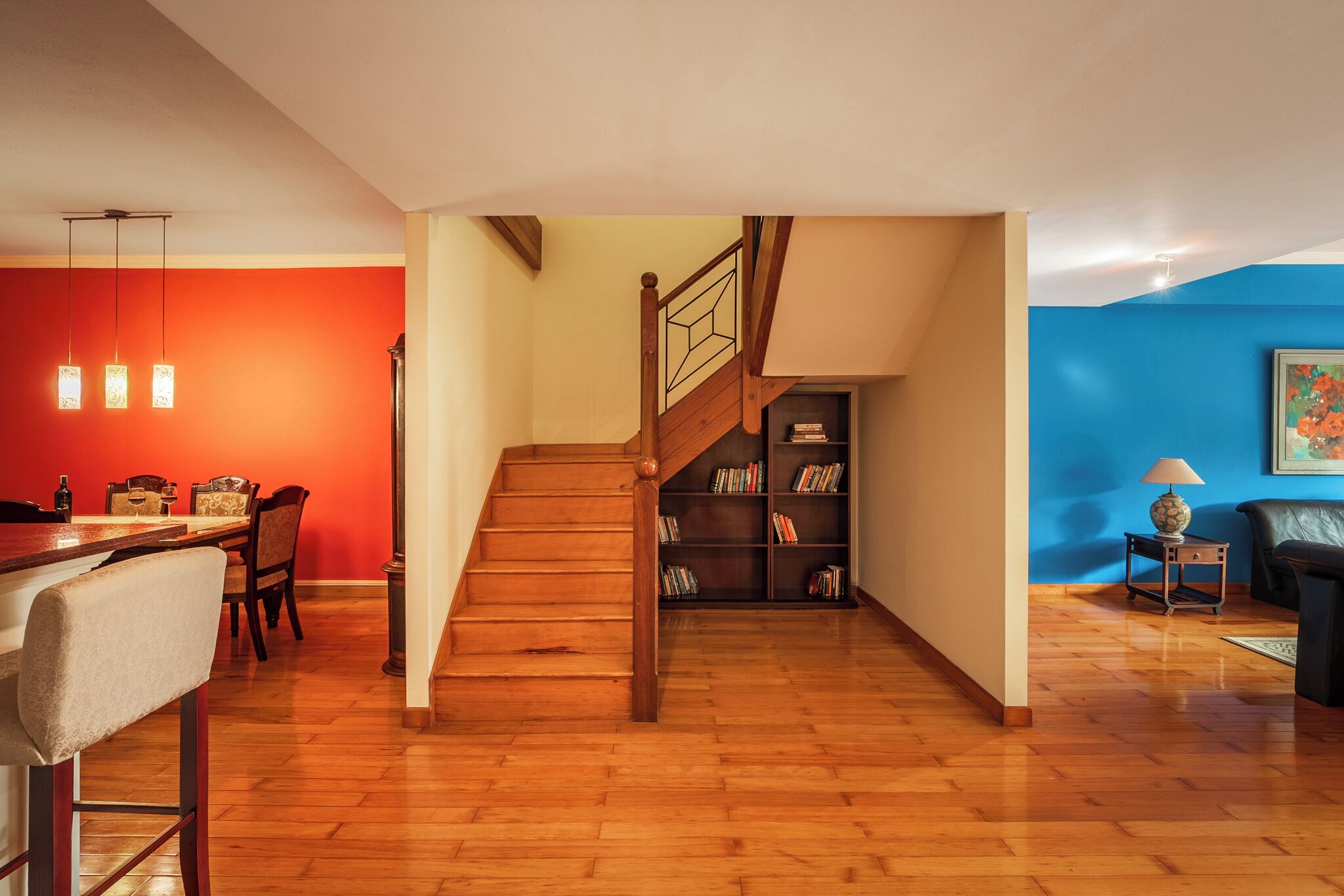 A modern living space with a wooden staircase, a dining area with a red wall, and a bookshelf.