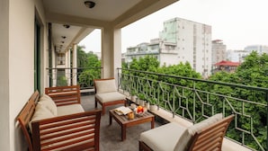 Un balcón con mobiliario de madera, una mesa de centro con frutas y un libro, y vistas a edificios de la ciudad.