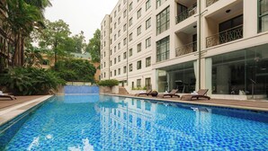 Una piscina con suelo de baldosas, rodeada de sillas de descanso y un edificio con varios balcones.