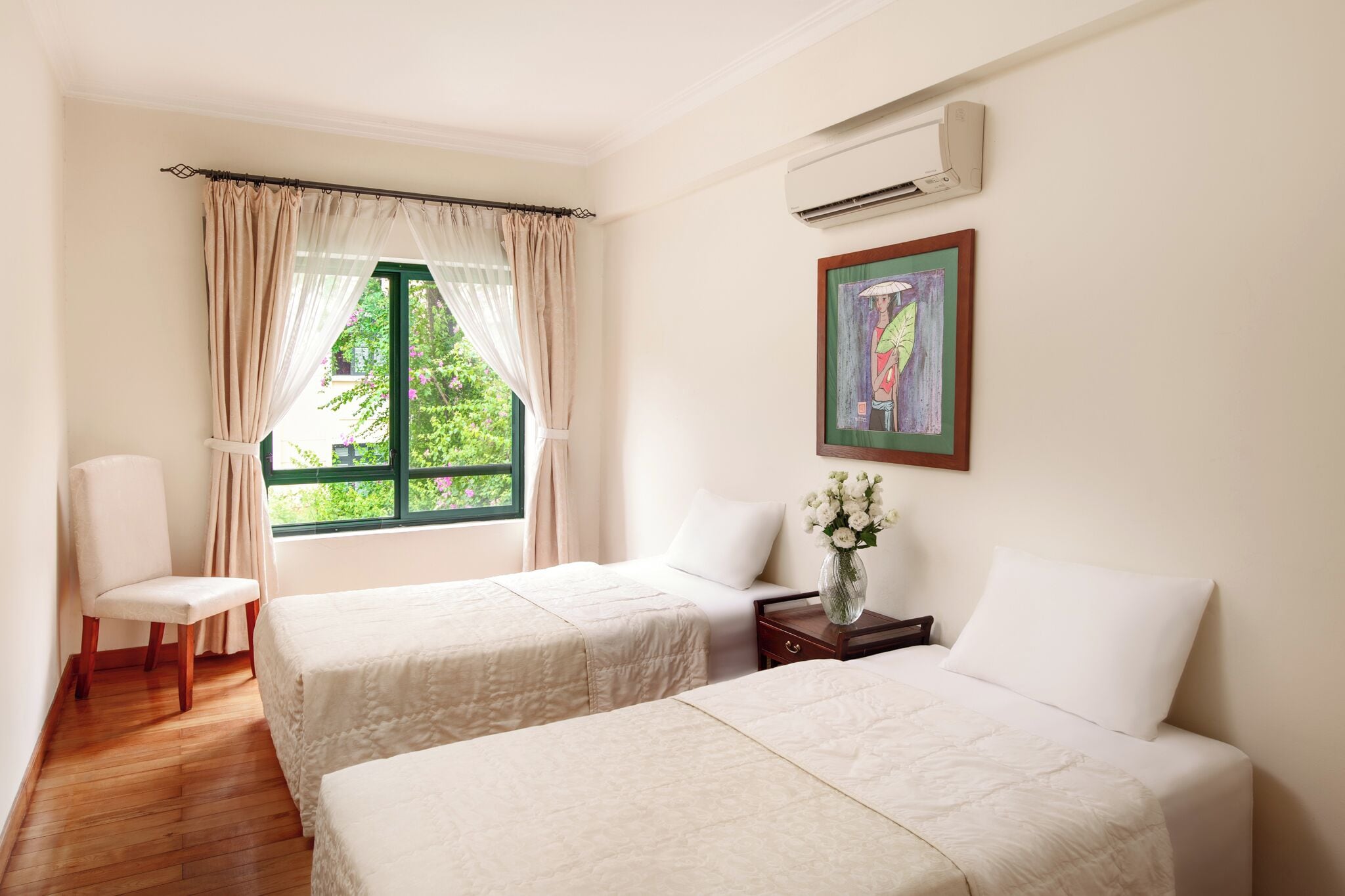 Quarto de hotel com duas camas, uma cadeira, uma mesa de cabeceira, um vaso com flores e uma janela com cortinas.