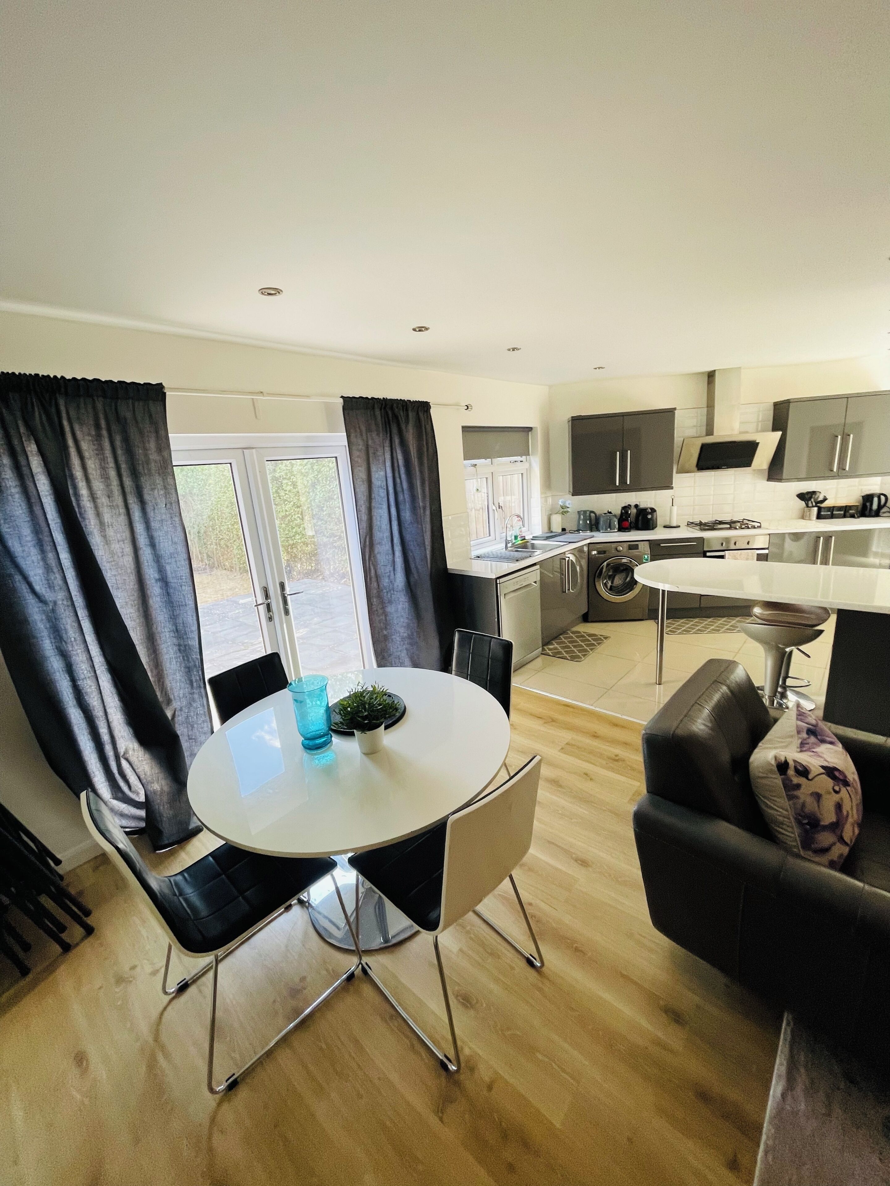 A modern kitchen with a dining area, including a round table with chairs, a sofa, and a window with curtains.