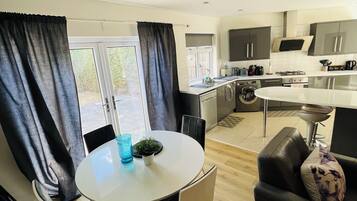 A modern kitchen with a dining area, including a round table with chairs, a sofa, and a window with curtains.