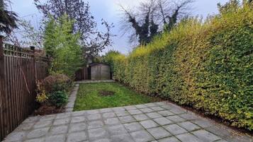 A paved garden pathway leading to a grassy area with a wooden shed and a tall hedge.