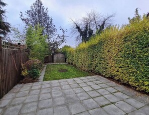 Un sendero pavimentado en un jardín que conduce a una zona de césped, con un cobertizo de madera y un seto alto.
