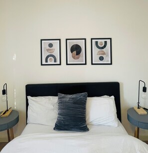 A bed with white bedding and a dark headboard, flanked by two bedside tables with lamps. Three framed abstract artworks are hung on the wall above the bed. - Spacious House | Sleeps 10 | 15mins to LDNcentre | 3 Private Parking Spots (London)