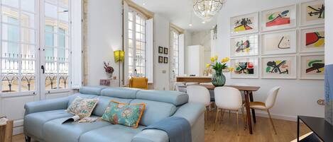 A modern living room with a light blue sofa, a chandelier, and a dining area with white chairs and a wooden table.