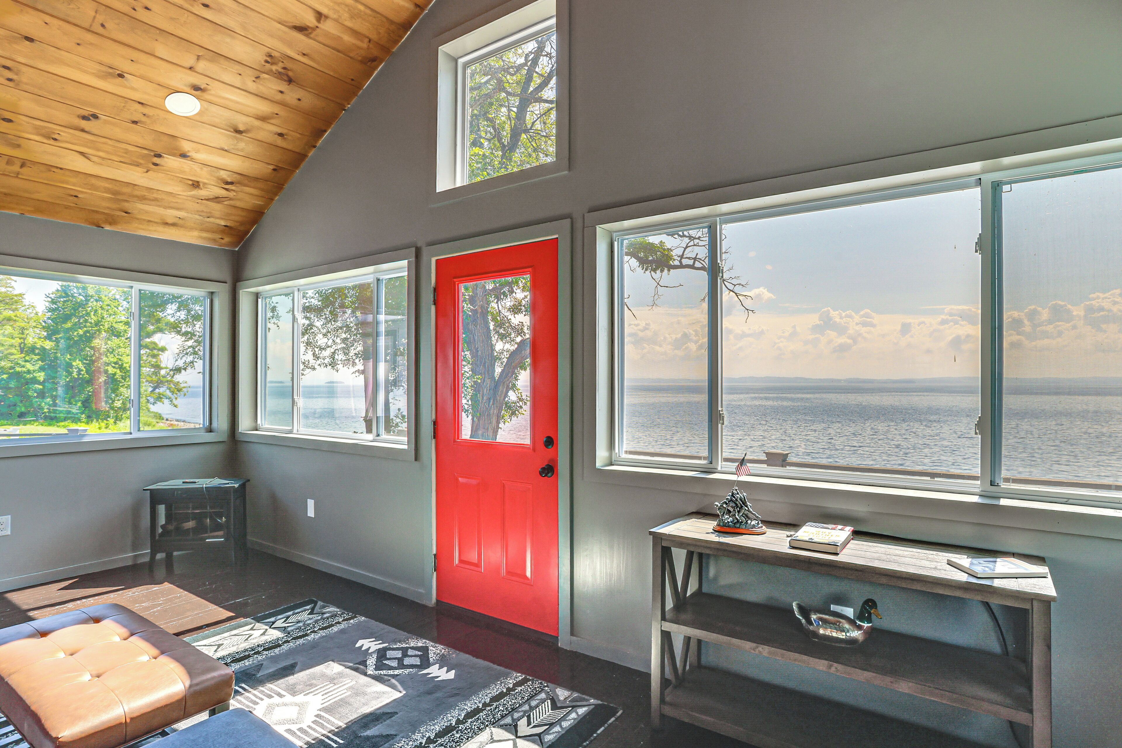 A room with a red door, large windows, a wooden ceiling, and a view of the water.