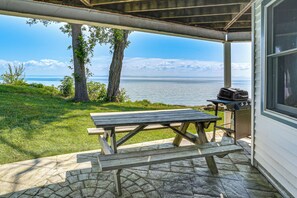 En täckt uteplats med ett picknickbord och en grill, med utsikt över en vattenyta och grönområden.