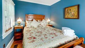 Ein Schlafzimmer mit floralem Bettüberwurf, Holz-Kopfschild, zwei Nachttischlampen und einem gerahmten Blumenbild an der Wand.