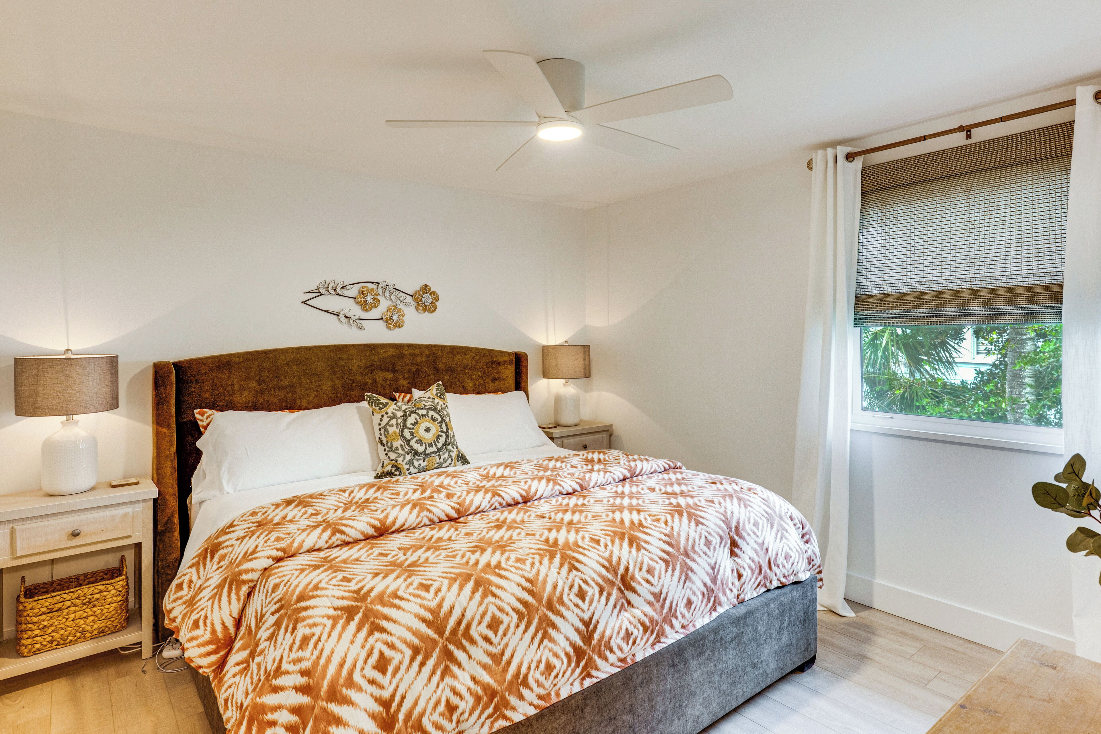 Ein Schlafzimmer mit einem Bett, einem Deckenventilator, einem Nachttisch, einer Lampe und einem Fenster mit Blick auf Grün.