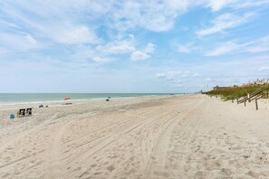Ein weiter Sandstrand mit blauem Himmel, vereinzelten Sonnenschirmen und einem Holzzaun entlang der Küste.