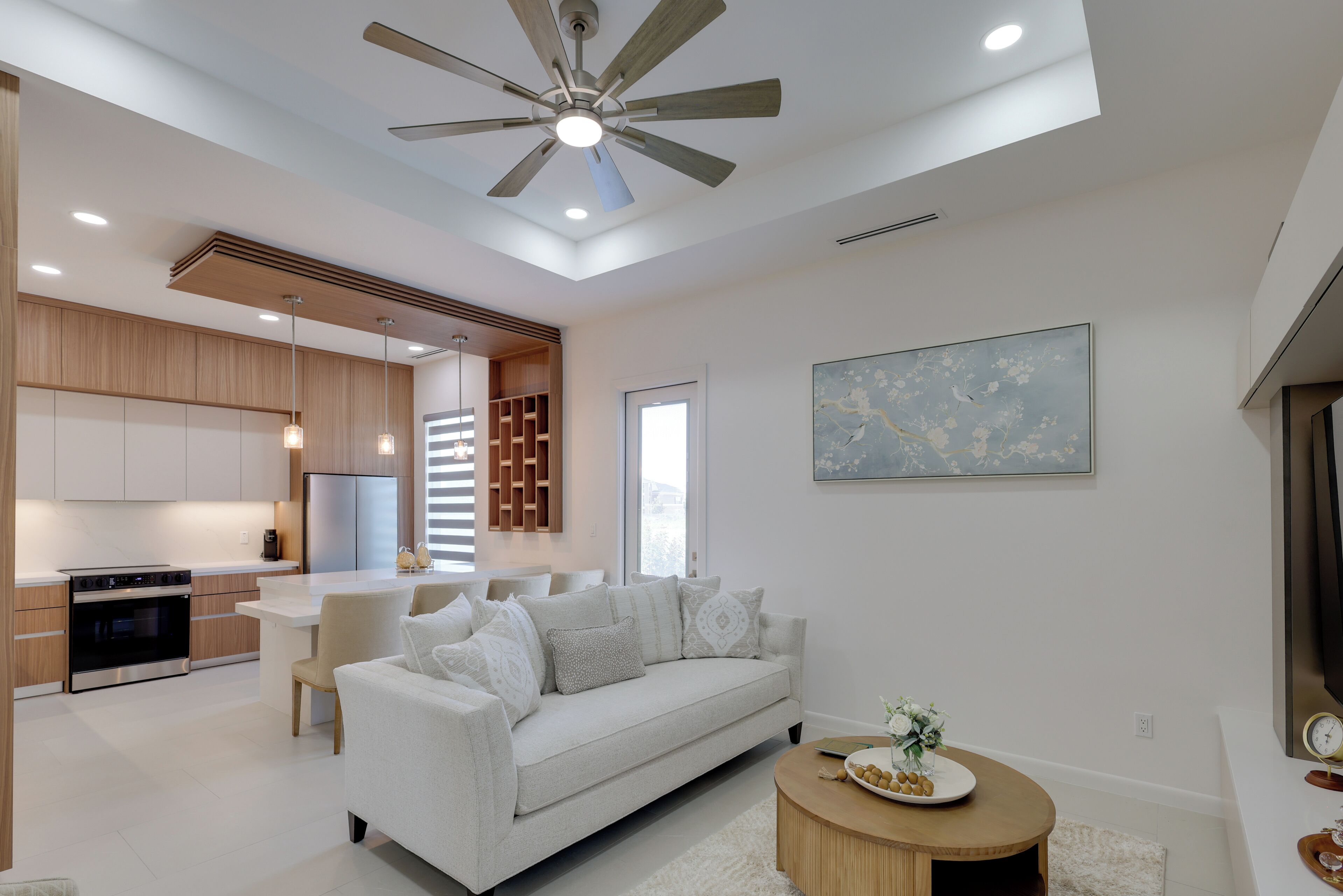 A modern kitchen with built-in appliances, a dining area with a white table and chairs, a living room with a sofa and decorative pillows, and a ceiling fan.