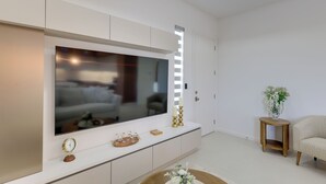 Ein modernes Wohnzimmer mit einem großen Flachbildfernseher an einem weißen Schrank, einem kleinen Holztisch mit einer Blumenvase und einem beige farbenen Sofa.