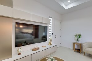 Ein modernes Wohnzimmer mit einem großen Flachbildfernseher an einem weißen Schrank, einem kleinen Holztisch mit einer Blumenvase und einem beige farbenen Sofa.