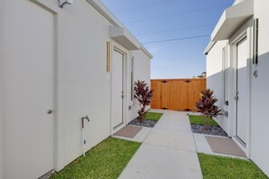 A narrow pathway leading to a wooden gate, flanked by white buildings with doors and windows, and small plants in the ground.