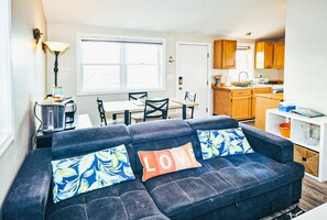 Sala de estar con sofá, dos sillones, una mesa de centro y una lámpara. Se observa una cocina con armarios de madera y estufa.
