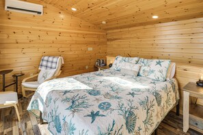 A wooden cabin bedroom with a bed, a chair, a small table, and an air conditioning unit.