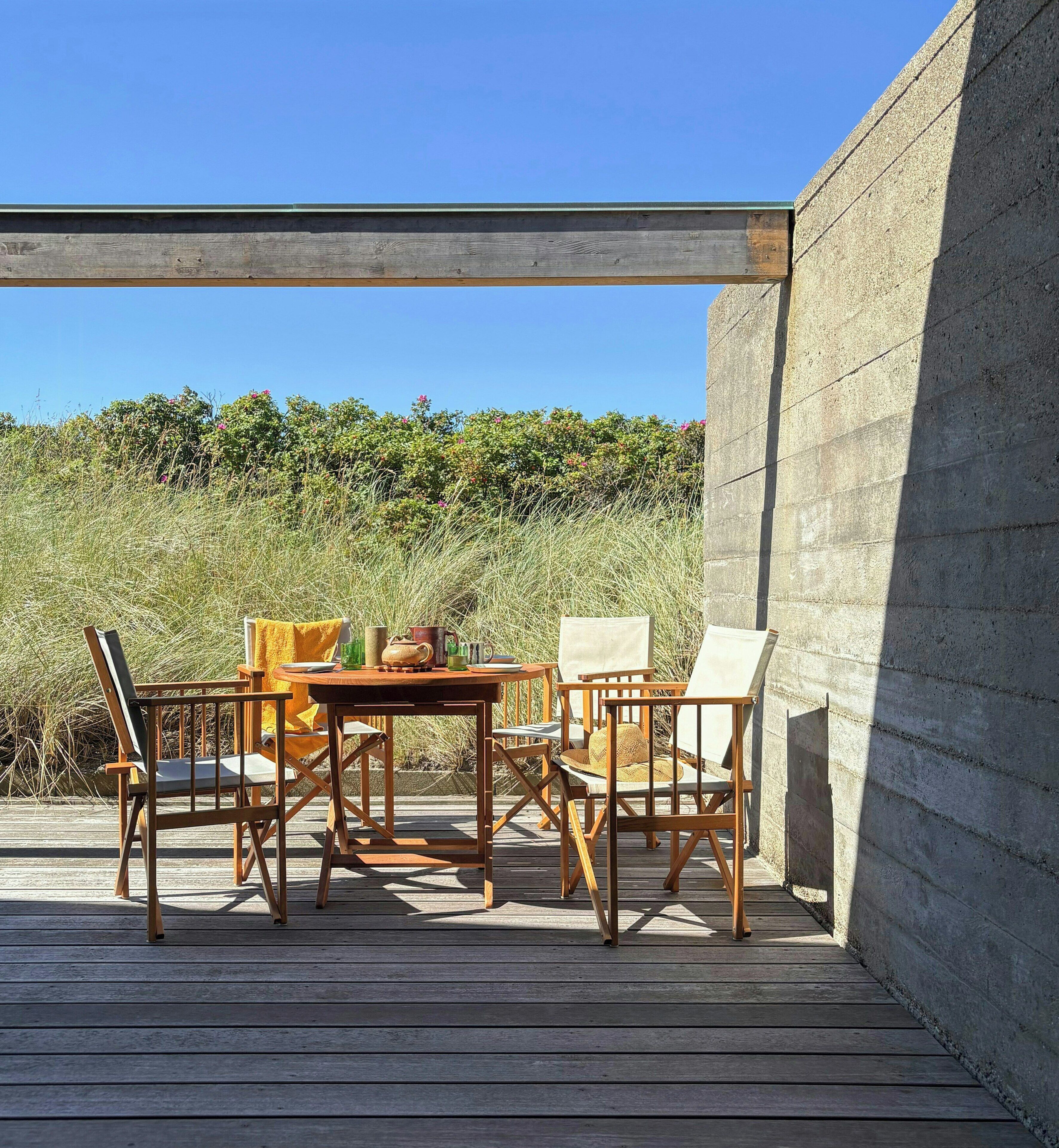 Eine Terrasse aus Holz mit Tisch und Stühlen, eine Betonmauer und Blick auf Strand und Vegetation.
