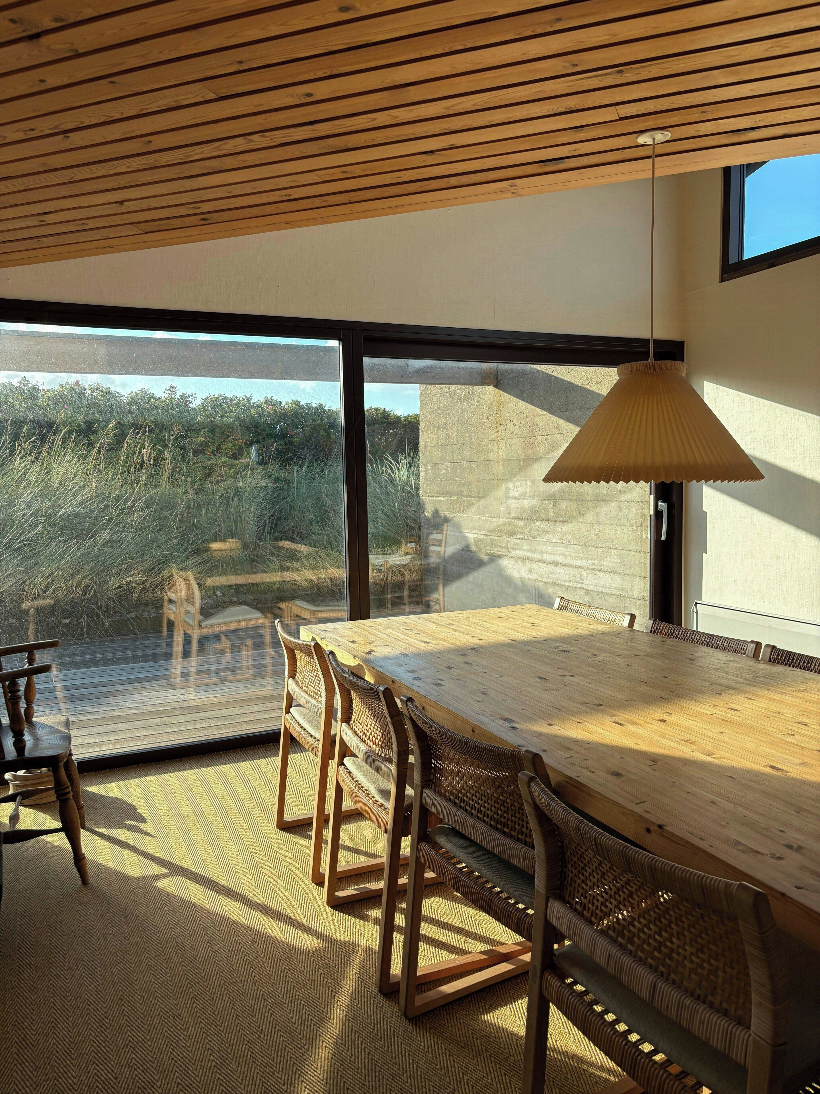 Comedor con mesa y sillas de madera, ventanas amplias y una lámpara colgante.
