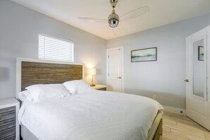 Ein Schlafzimmer mit Bett, Nachttisch, Lampe, Deckenventilator, Fenster mit Jalousien, einer Tür und einem Bild an der Wand.