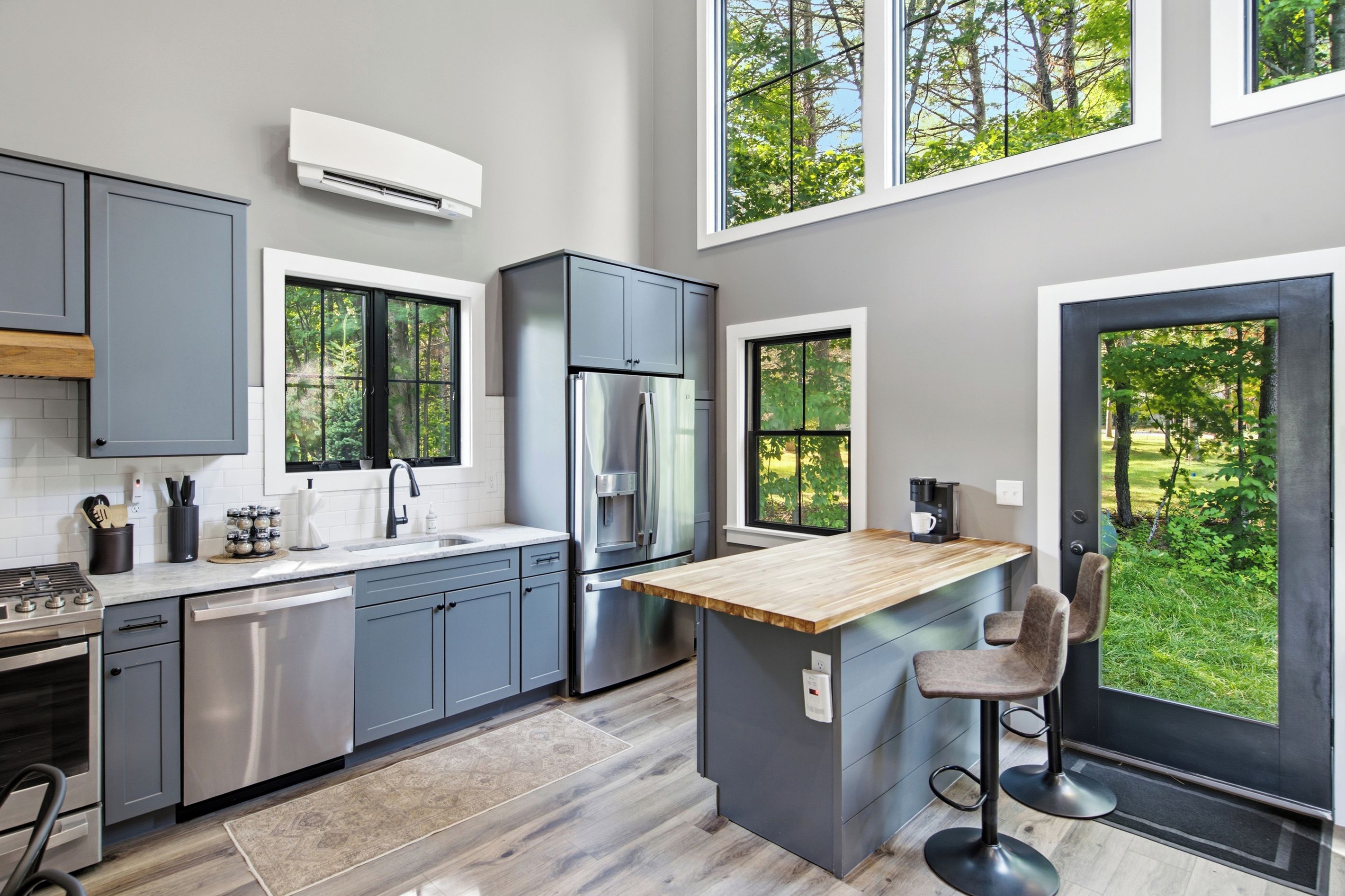 A modern kitchen with grey cabinets, stainless steel appliances, a wooden island, and large windows offering views of greenery.