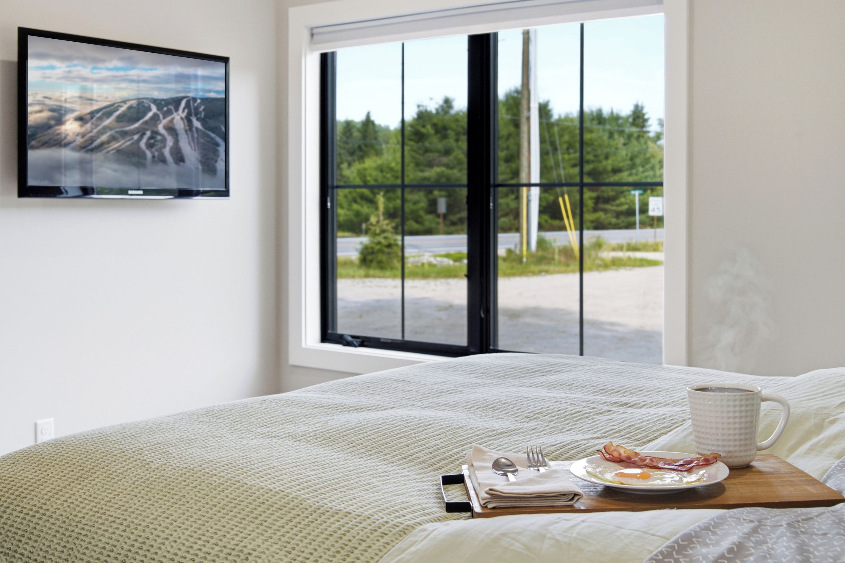 A neatly made bed with a breakfast tray on it, a window with a view of greenery, and a framed picture on the wall.