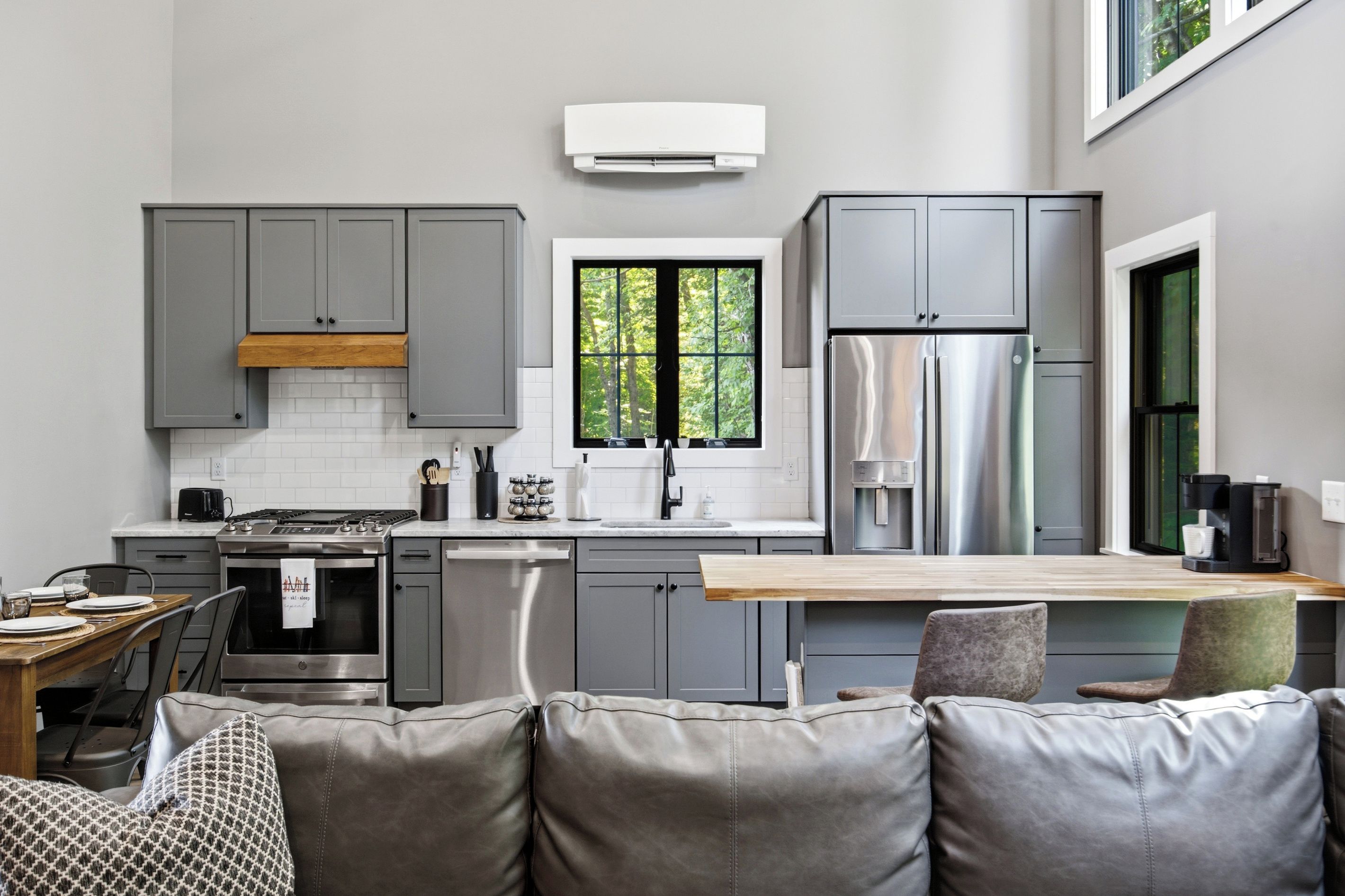 A modern kitchen with grey cabinets, a stainless steel refrigerator, and a window with a view of trees.