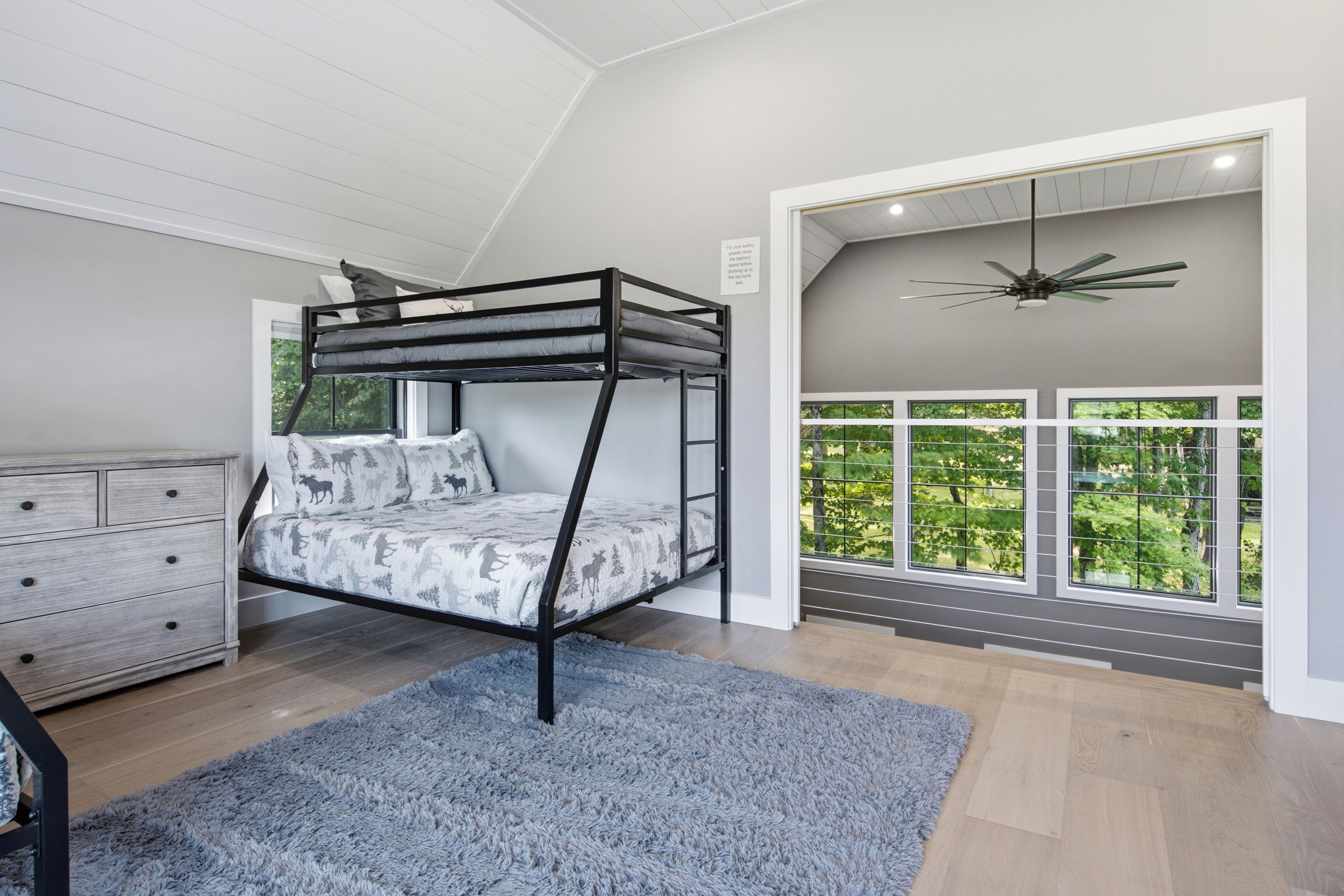 A bunk bed with a forest-themed bedding, a grey dresser, a blue rug, and a view of greenery through large windows.