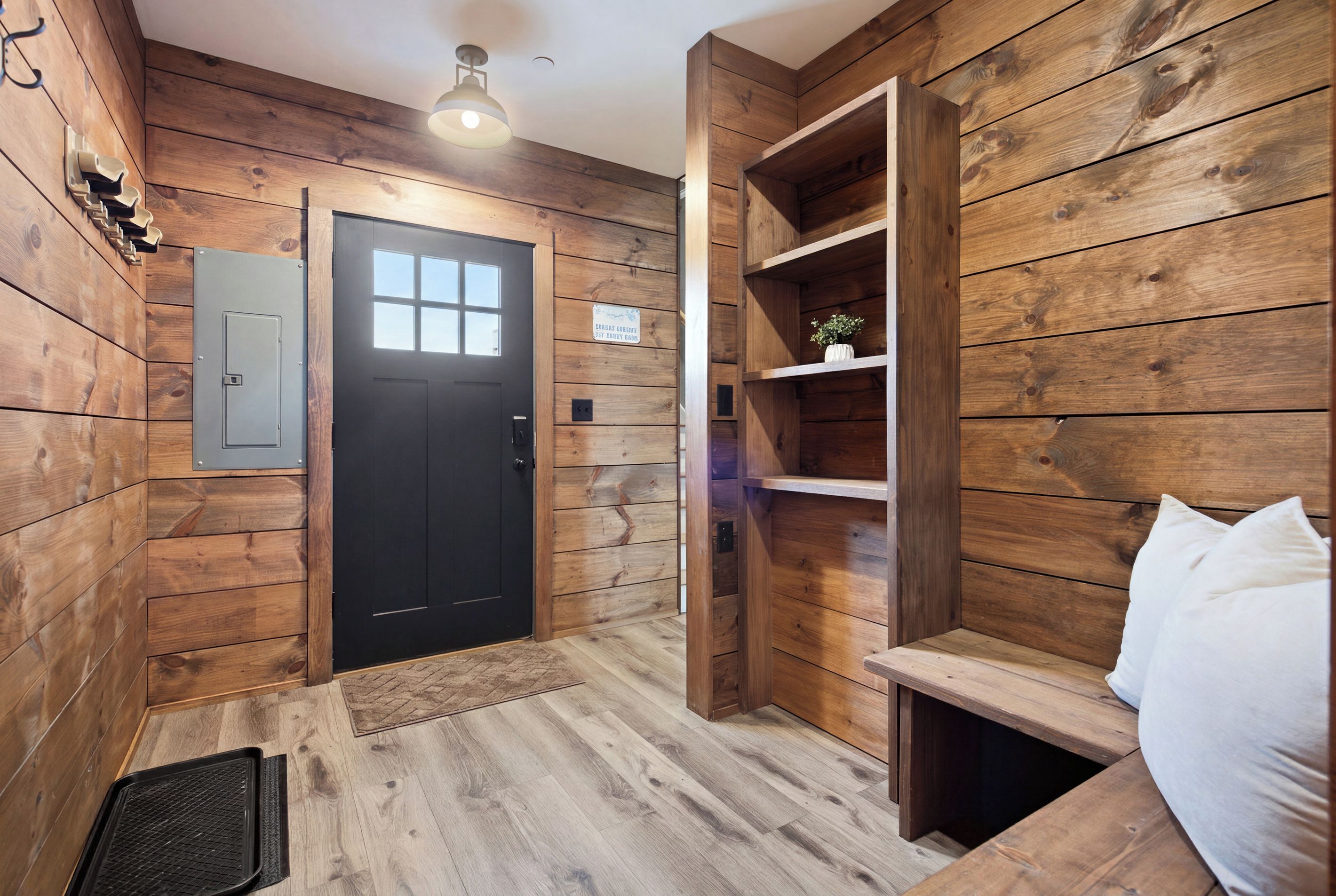 Un intérieur en bois avec une porte noire, une étagère ornée d’une plante et un coussin blanc.