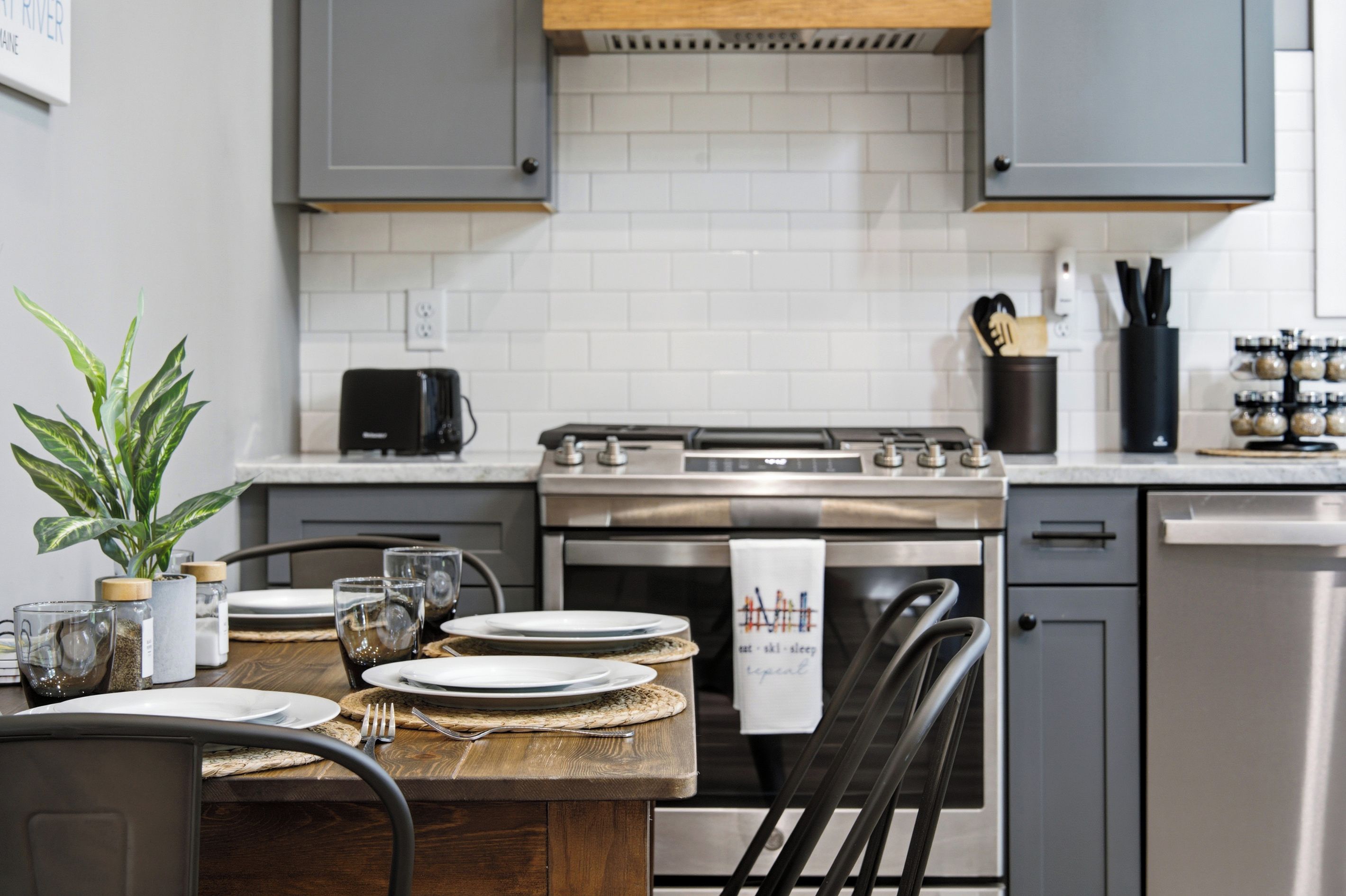 A modern kitchen with grey cabinets, a stainless steel stove, and a dining table set with plates and glasses.