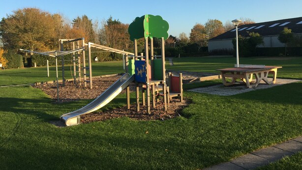 A playground with a slide and climbing structures, surrounded by grass and trees.