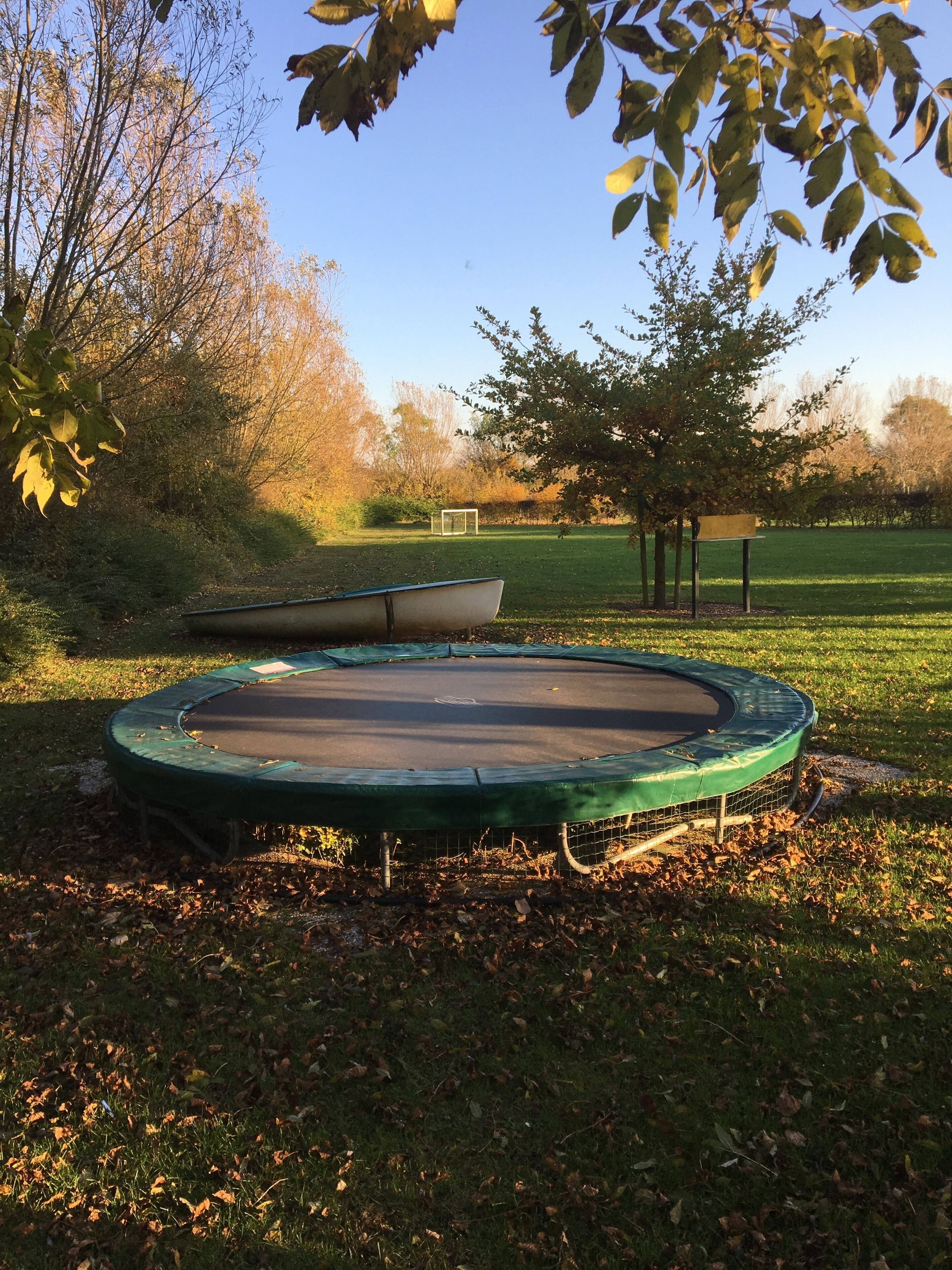 A trampoline in a grassy area with trees and a soccer goal in the background.
