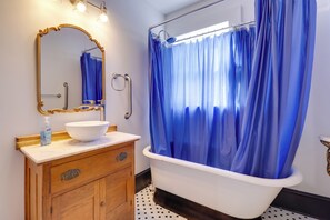 Ein Badezimmer mit einer weißen Badewanne, einem blauen Duschvorhang, einem hölzernen Waschtisch mit einer weißen Spüle und einem spiegel mit goldener Rahmung.