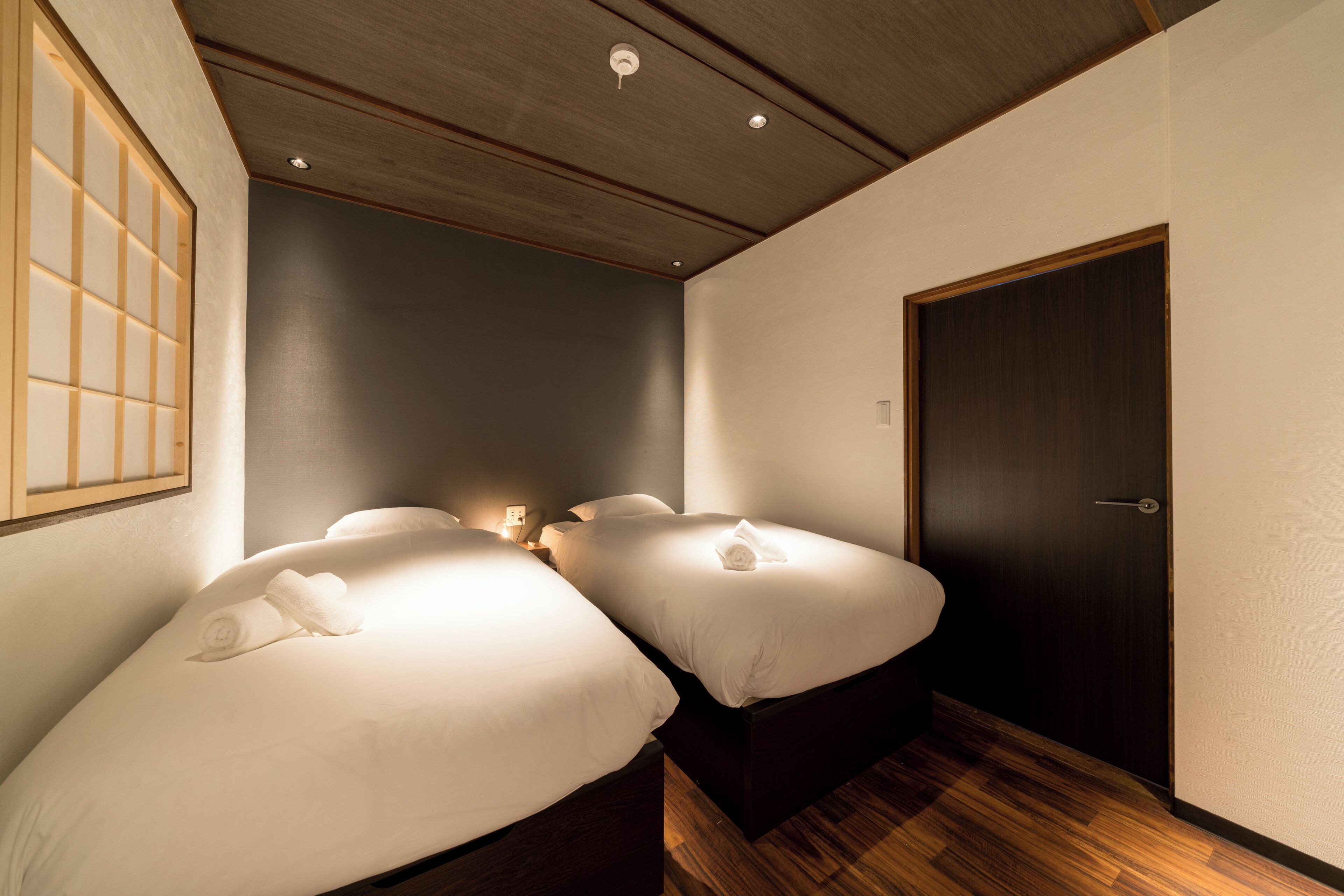 A hotel room with two beds, a wooden floor, a dark door, and a wall-mounted light fixture.