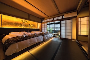 A traditional Japanese-style room with tatami flooring, a large bed with multiple pillows, a sliding shoji screen door, and a view of greenery outside. - Vrbo Property (大阪市東成区東小橋2丁目)
