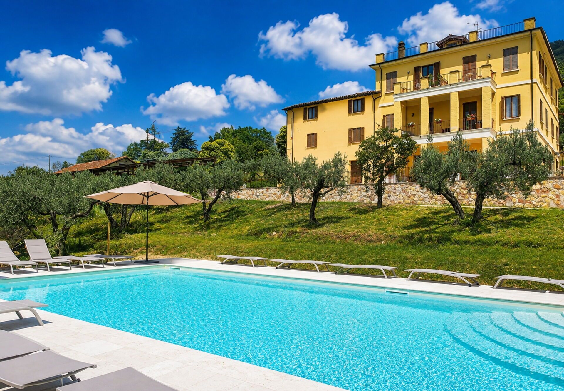 A swimming pool with lounge chairs and an umbrella, surrounded by a stone wall and greenery.