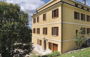 Ein mehrgeschossiges Gebäude mit gelber Fassade und braunen Fensterläden, mit einem zentralen Eingang und einer kleinen Grünfläche.
