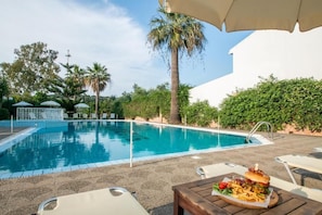 A swimming pool with lounge chairs and umbrellas, a table with food, and palm trees.