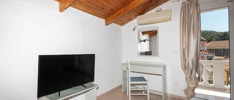 A room with a flat-screen TV on a stand, a desk with a chair, a window with a curtain, and an air conditioning unit on the wall.