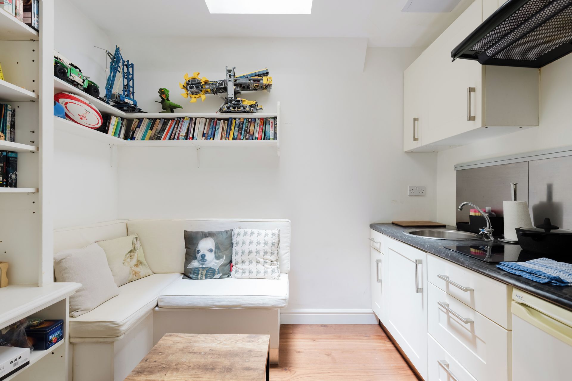 A compact living space with a kitchenette, a seating area with a sofa and pillows, and a bookshelf filled with books and toys.