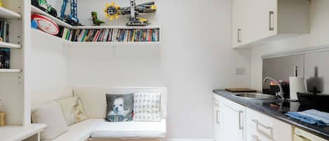 A compact living space with a kitchenette, a seating area with a sofa and pillows, and a bookshelf filled with books and toys.