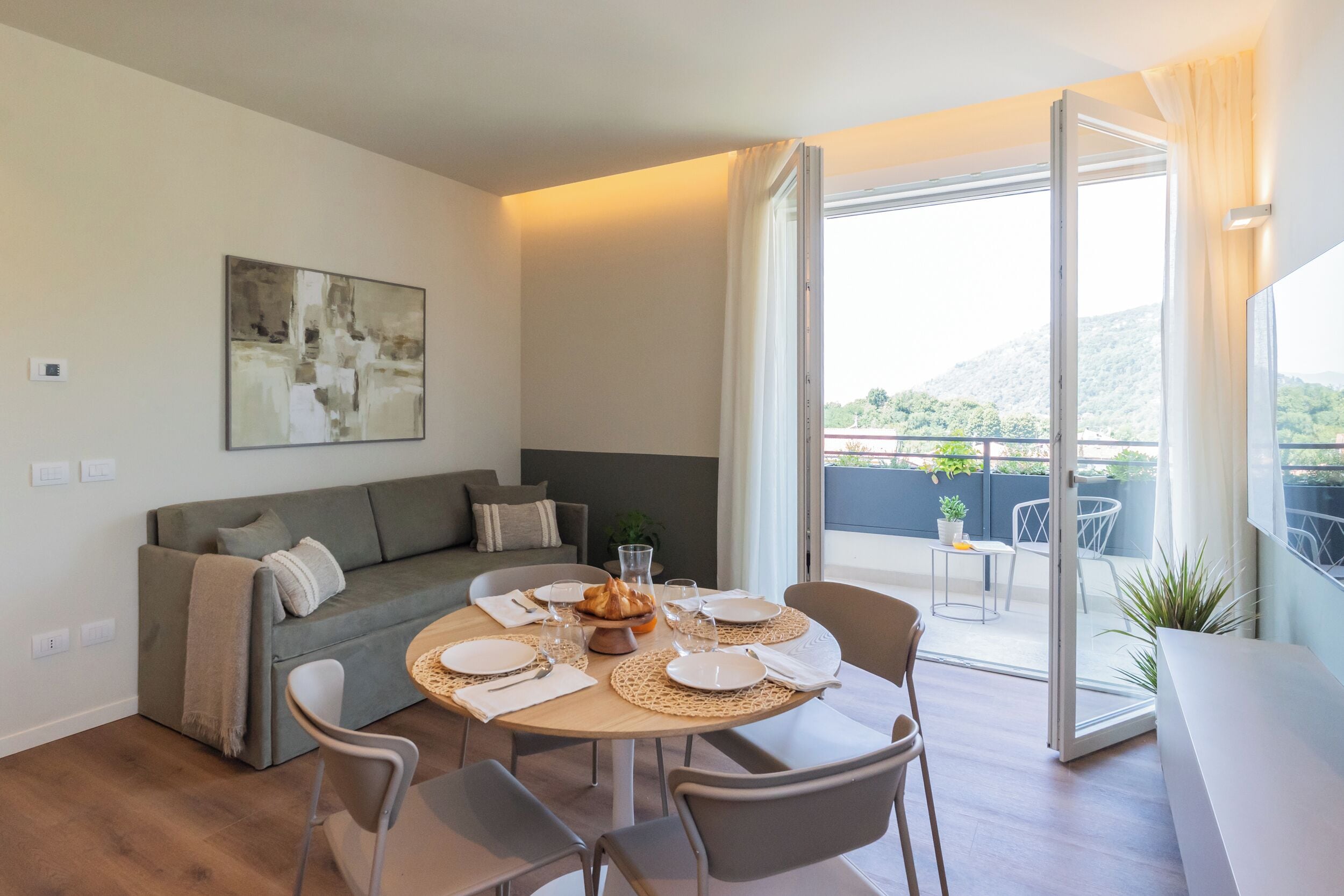 Un soggiorno moderno con zona pranzo, un divano, un tavolo apparecchiato per due e un'ampia porta-finestra scorrevole che dà su un balcone con vista su spazi verdi e colline.