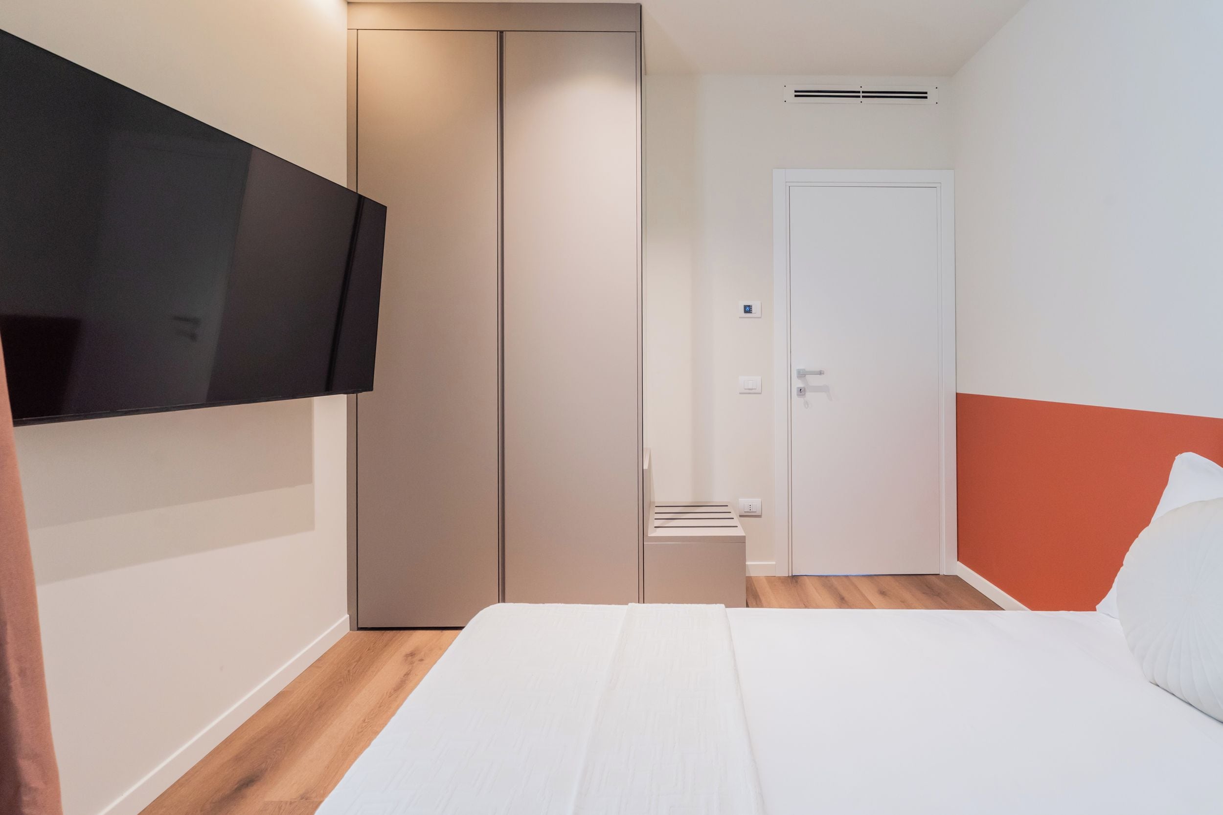 A bedroom with a bed, a wardrobe, a television mounted on the wall, and a door.