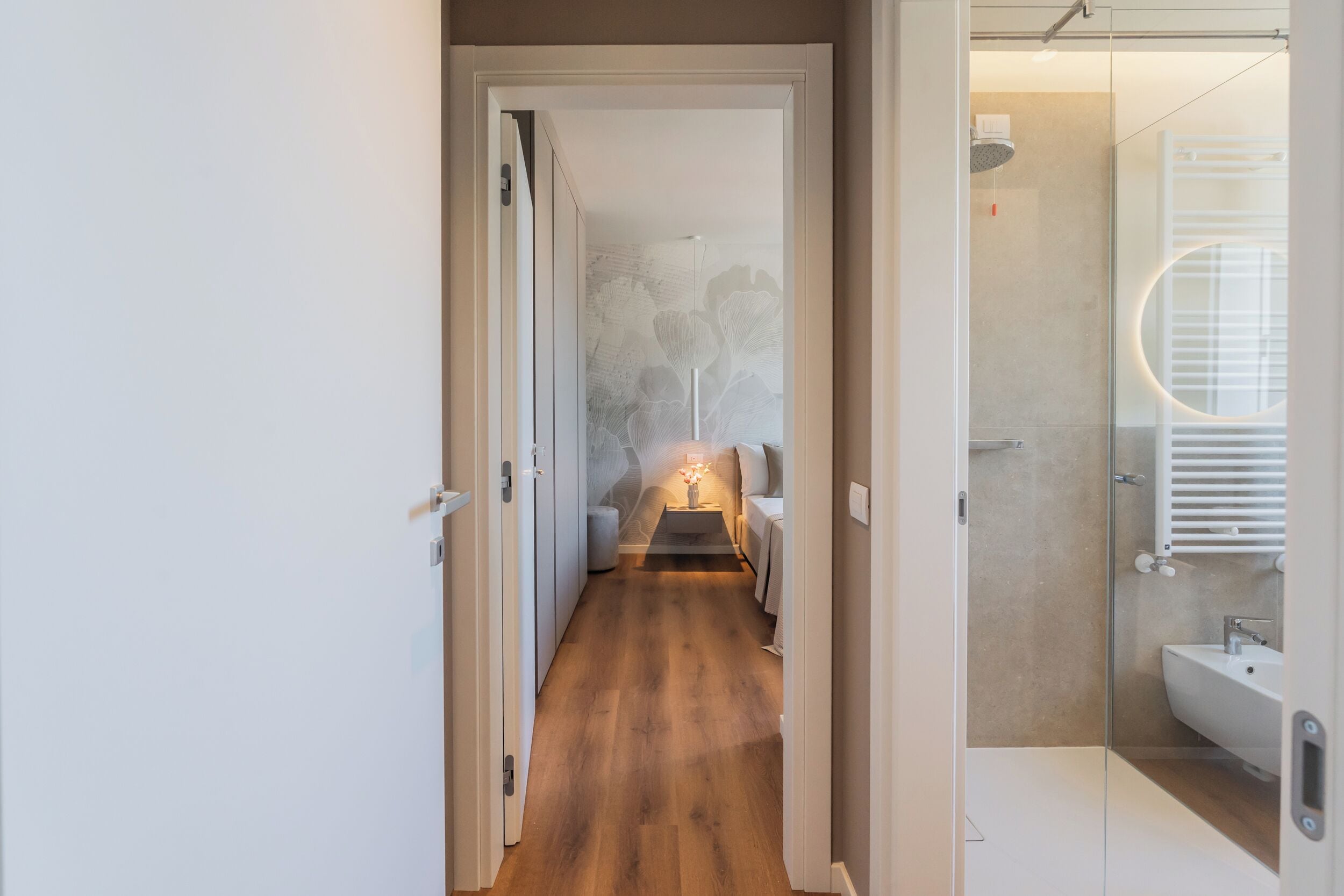 A hallway leading to a bedroom with a bed, bedside table, and a wall with a floral pattern.
