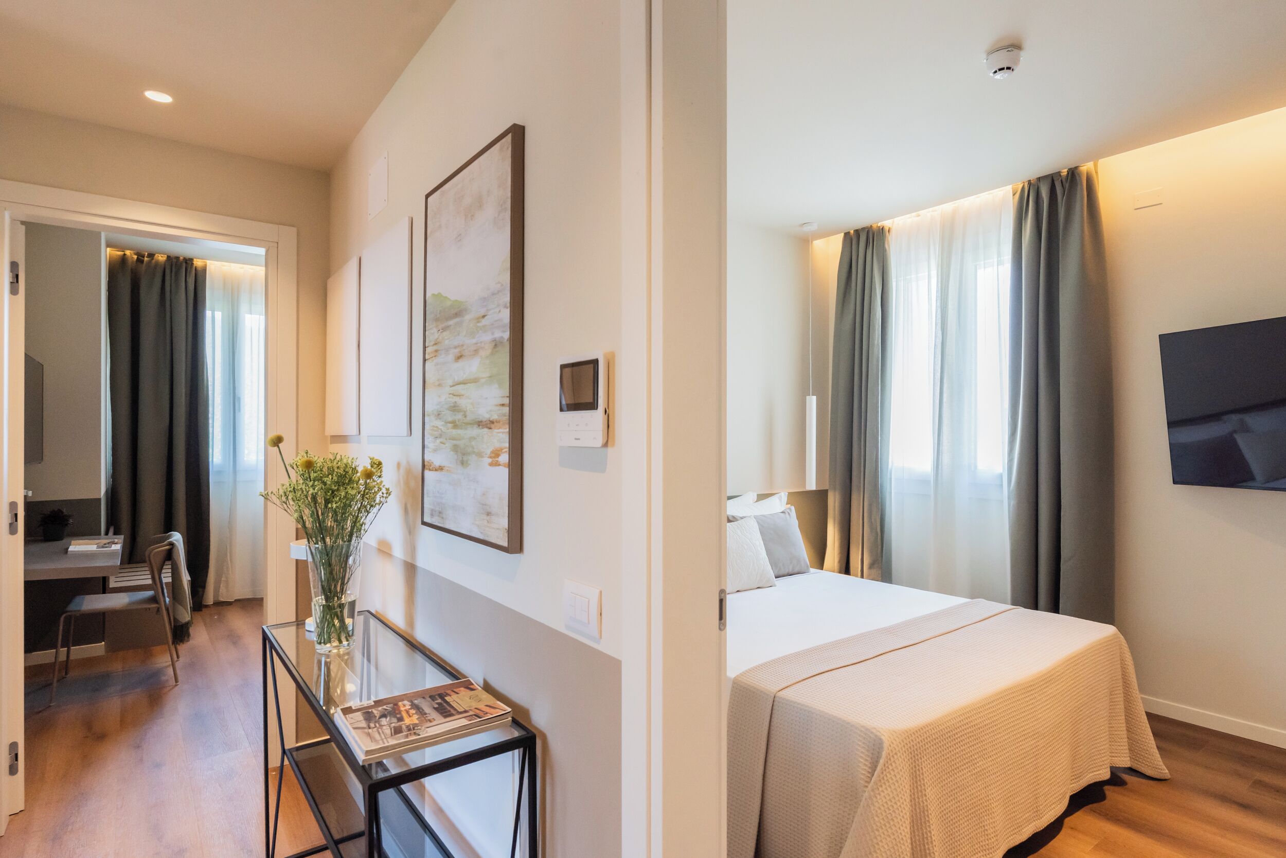Habitación de hotel con cama, escritorio, silla, una mesita con un florero y un cuadro en la pared.