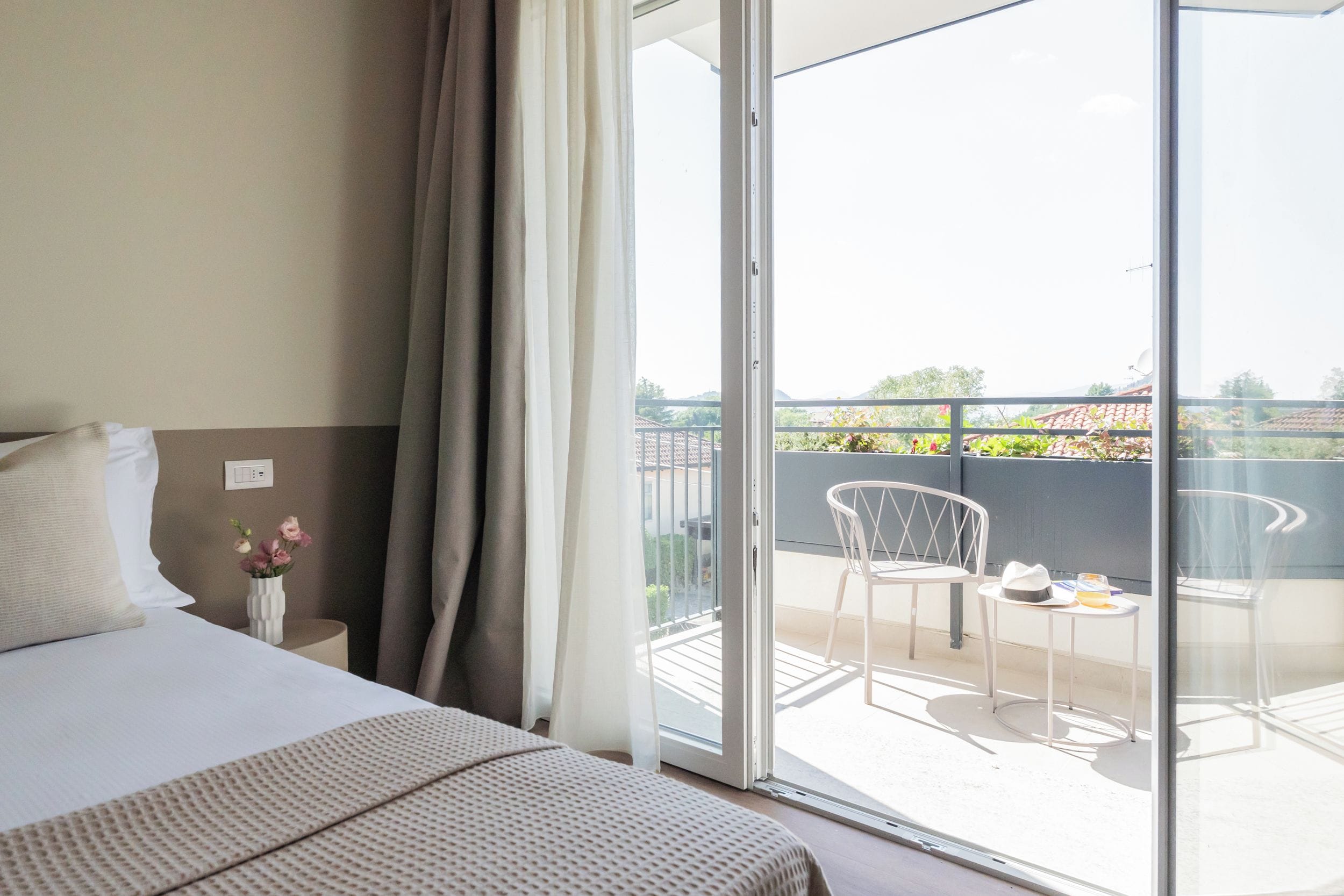 Una camera d'albergo con un letto, un comodino, un vaso di fiori e vista sul balcone esterno con sedie e un tavolo.
