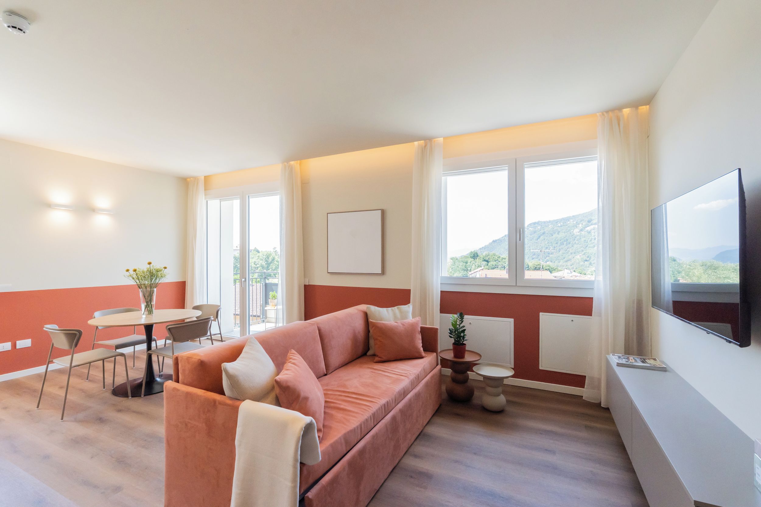 Una sala moderna con un sofá, una mesa de comedor con sillas, un televisor montado en la pared y vistas a zonas verdes.