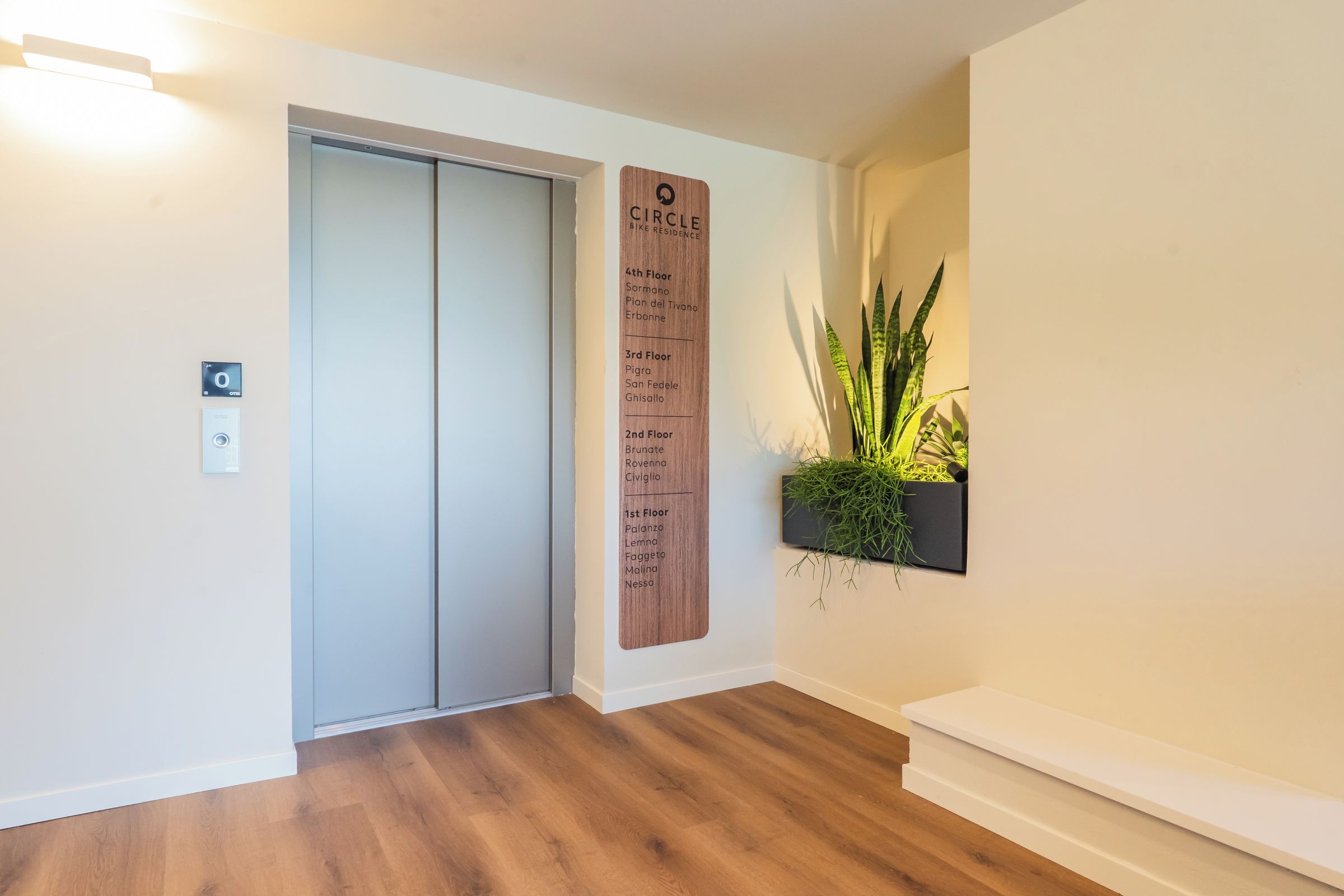 A hotel corridor with a wooden floor, a wall-mounted elevator, and a potted plant.
