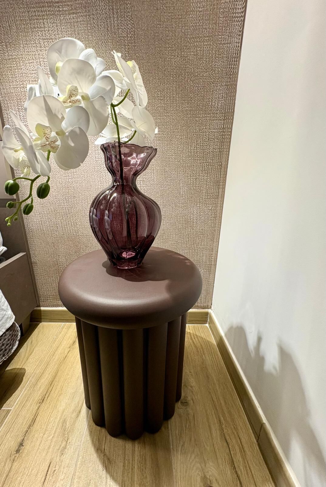 A corner of a room with a brown stool and a vase with white orchids.