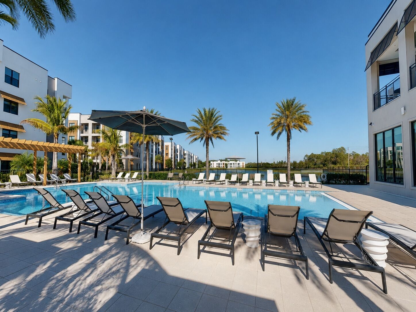 A pool area with lounge chairs and umbrellas, surrounded by palm trees and modern buildings.
